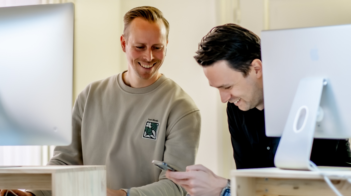 Twee mannen lachend achter een bureau met computers.
