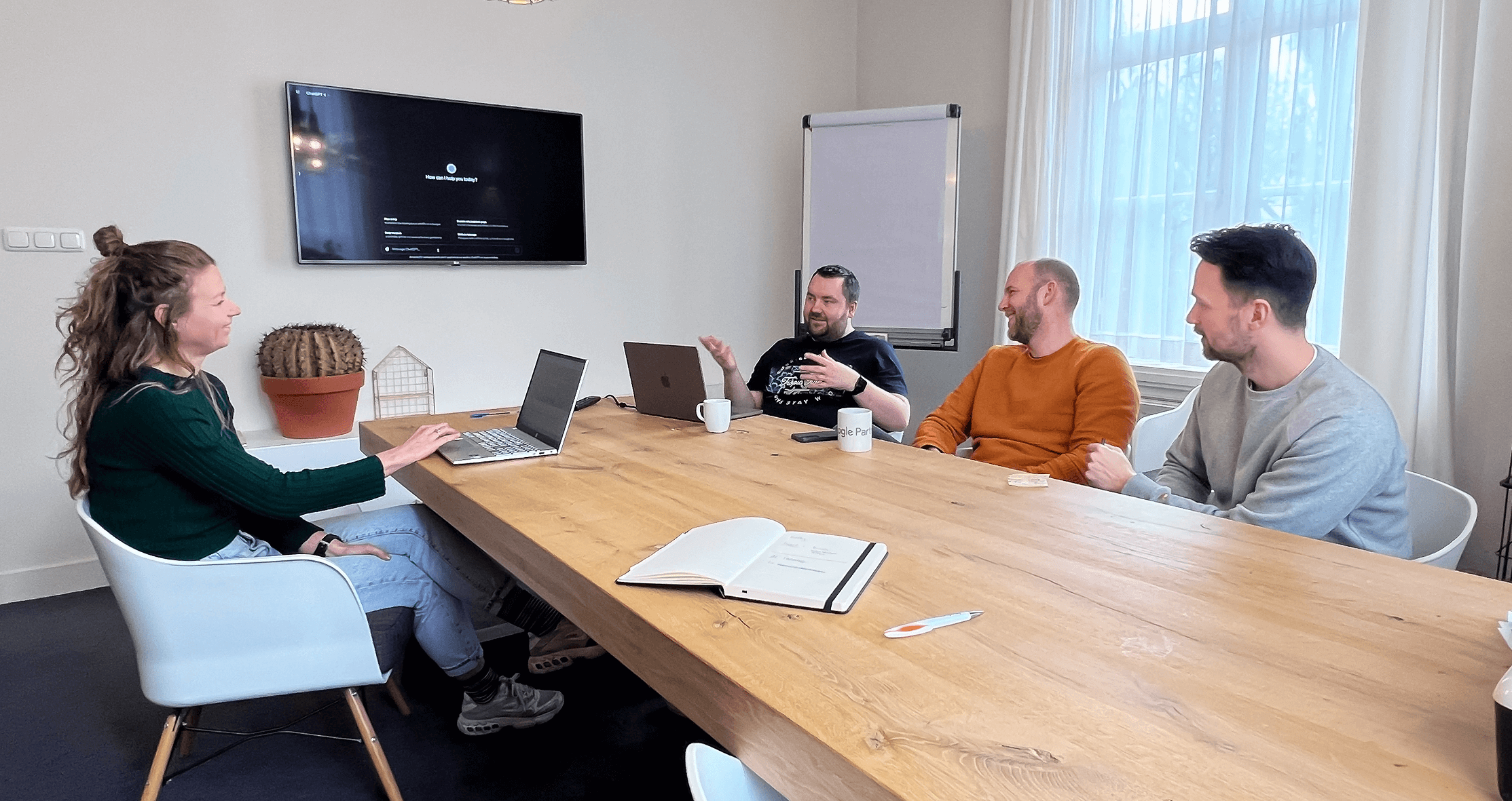 Vier mensen in een vergaderruimte met laptops en een scherm.
