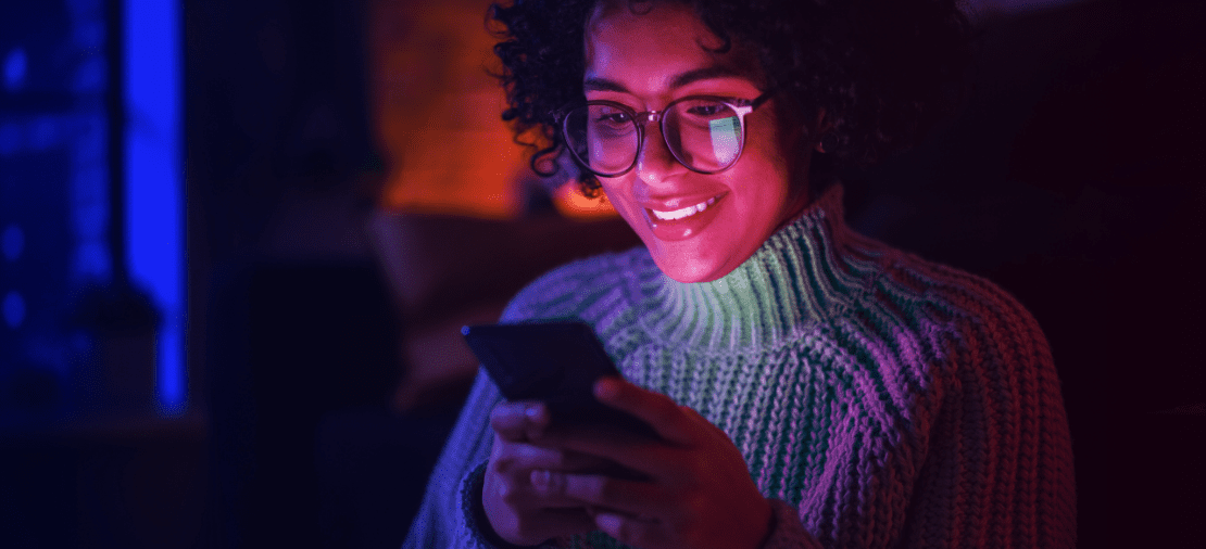 Student met bril kijkt lachend naar smartphone in donkere kamer.