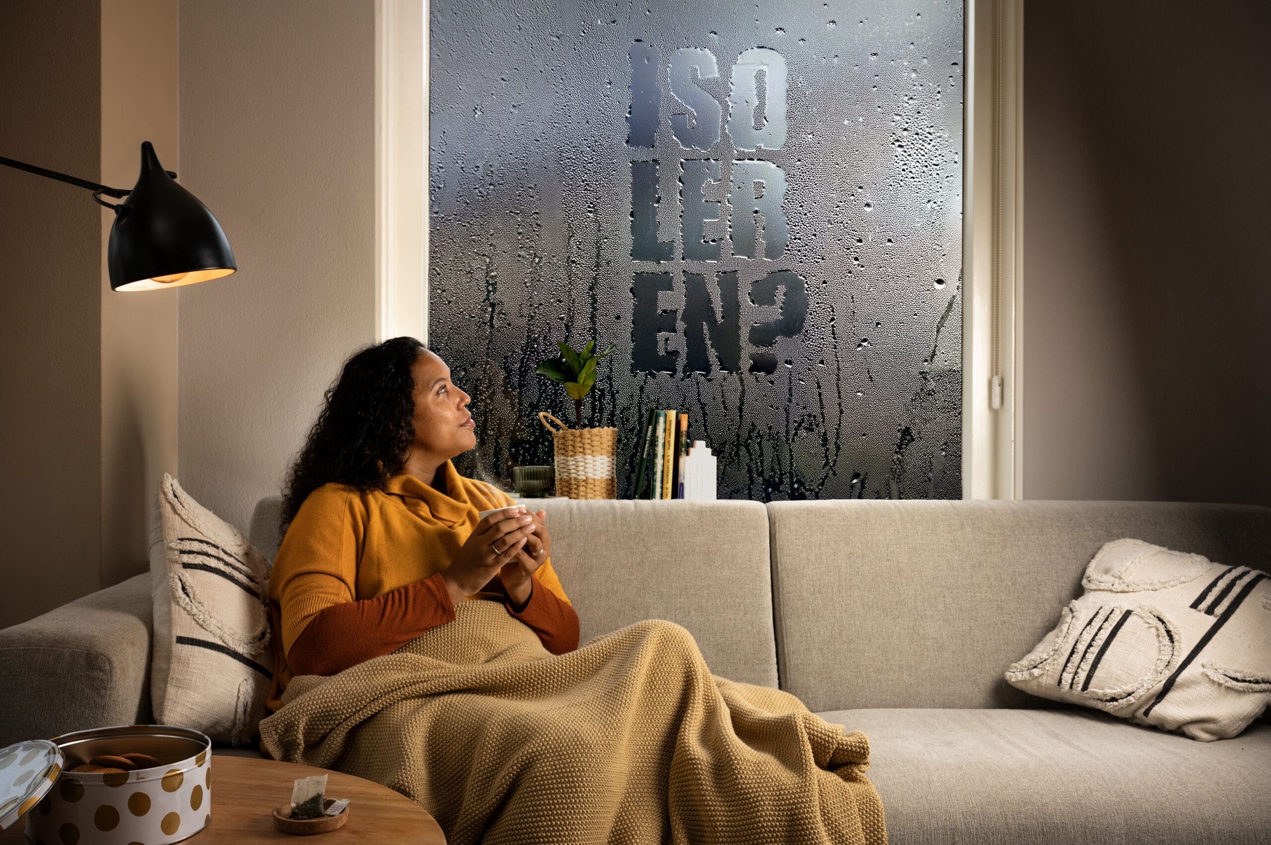 Vrouw zit met een deken en warme drank op de bank, kijkt naar een beslagen raam met de tekst 'Isoleren?' erin.