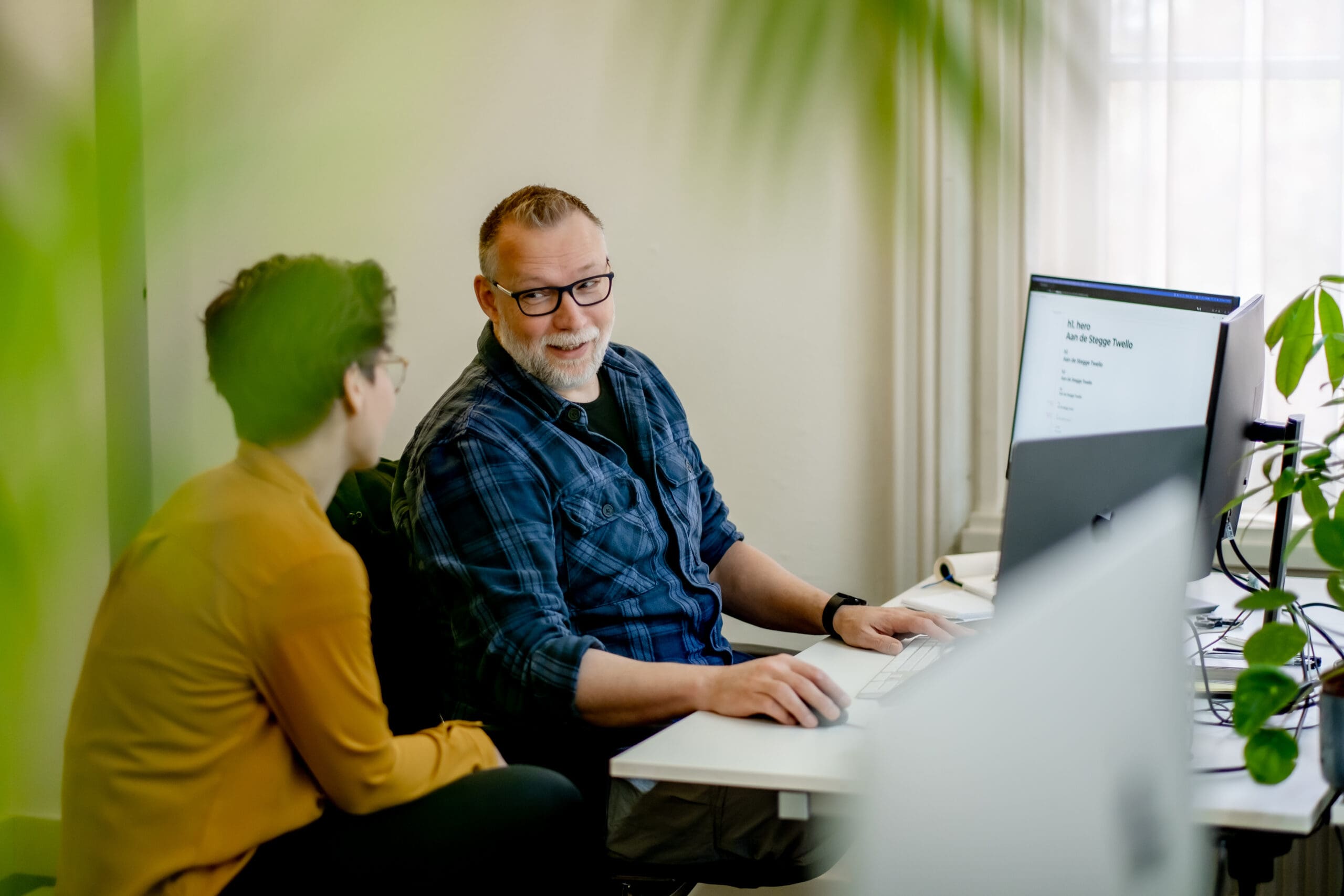 Twee collega’s in gesprek achter een bureau met computer in een informele kantooromgeving.