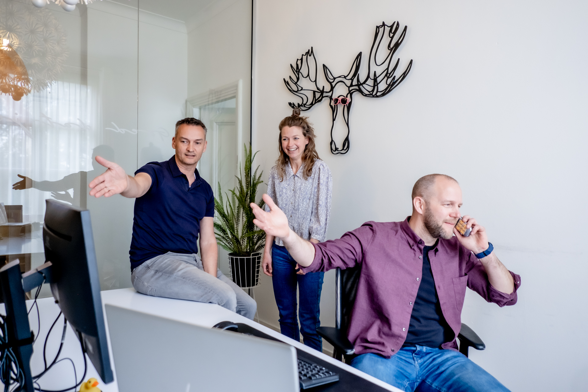 Drie collega’s op een kantoor; één wijst naar een computerscherm, een ander lacht en de derde telefoneert.